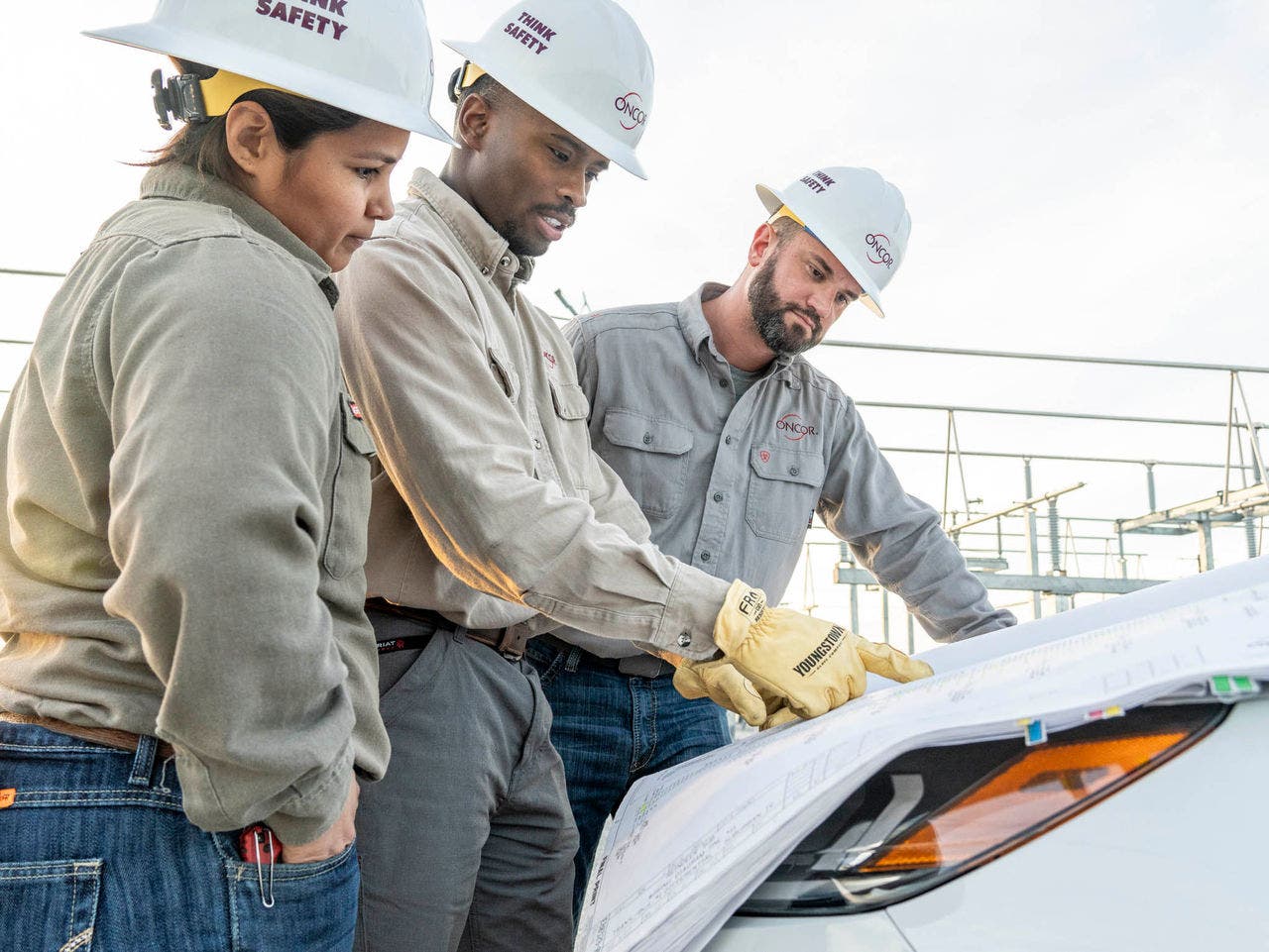 Three Oncor employees with hardhats study a document that is on the hood a car.